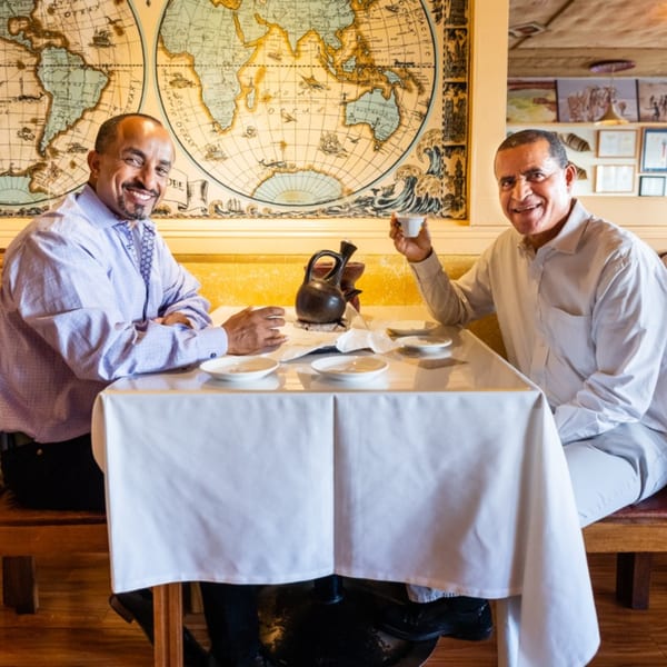 Two smiling men enjoying coffee at a table with a large world map on the wall.