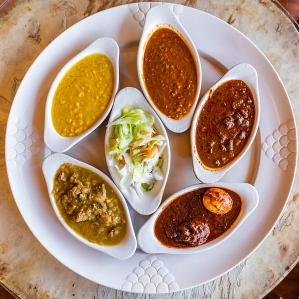 Overhead view of a white platter with six small bowls of colorful Ethiopian stews and salad.