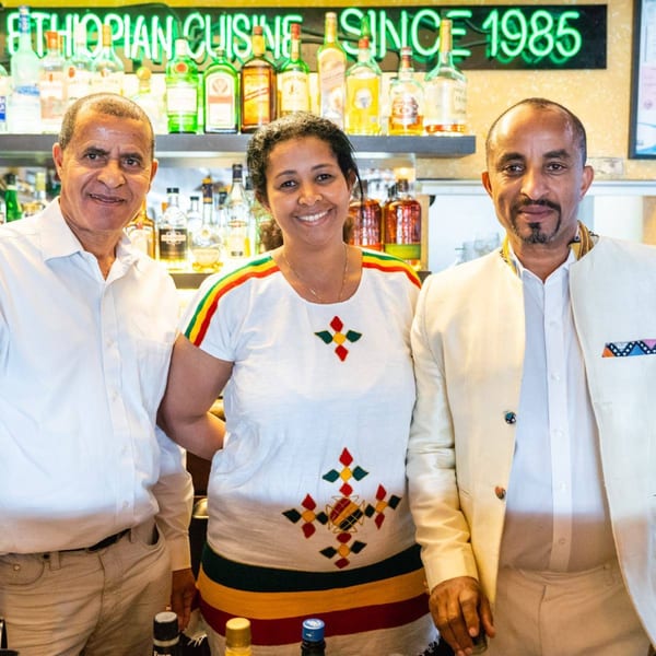 Three smiling Ethiopian restaurant staff, two men and one woman, stand in front of a bar.