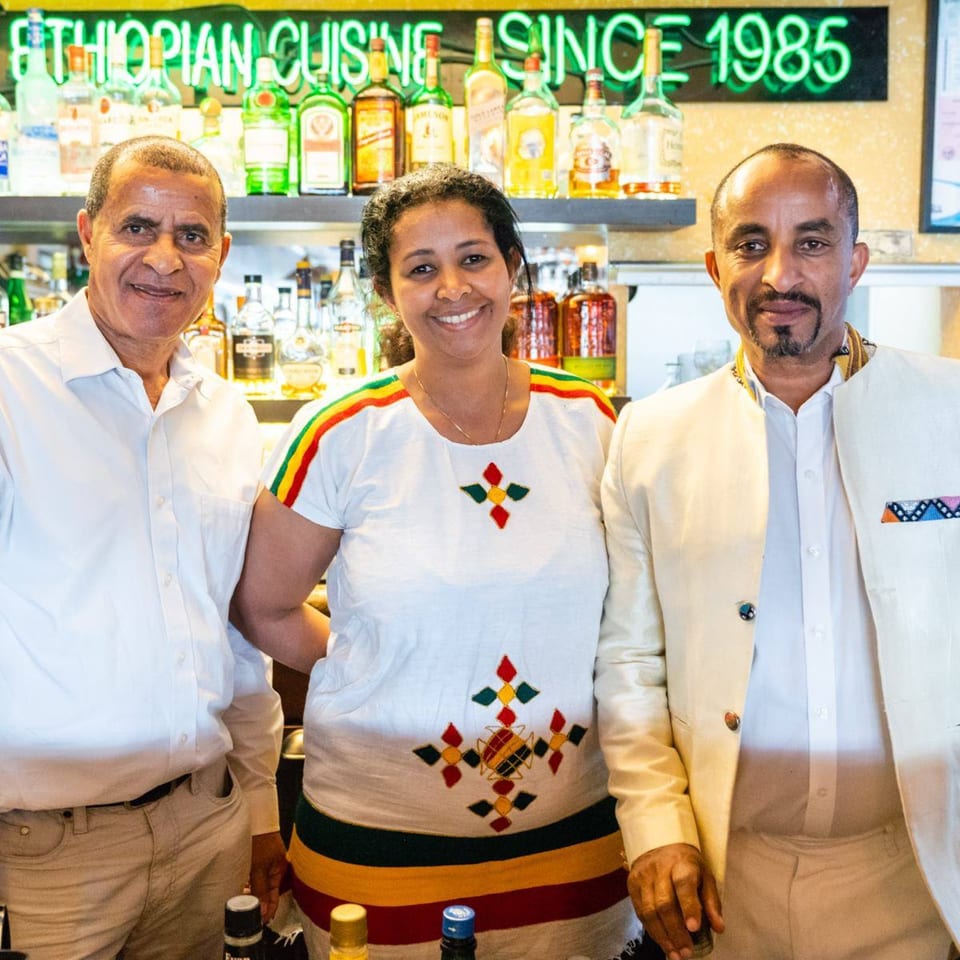 Three smiling Ethiopian restaurant staff, two men and one woman, stand in front of a bar.
