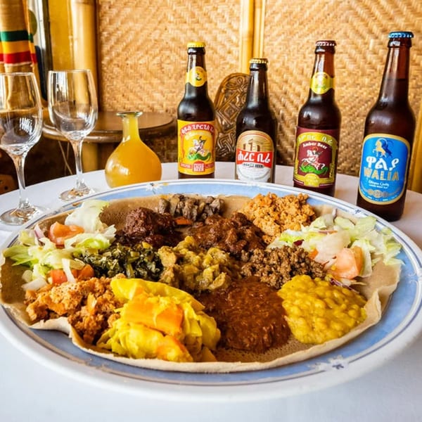 A large platter of Ethiopian injera bread topped with various stews, vegetables, and meats, with four bottles of beer and two wine glasses in the background.