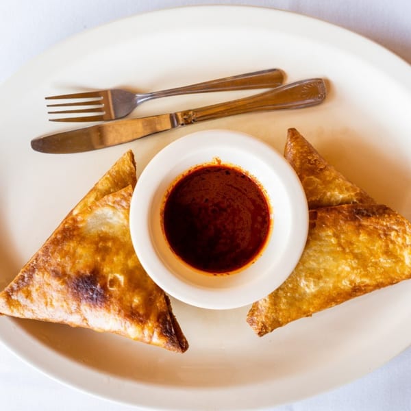 Two golden-brown Ethiopian sambusas flank a white bowl of red dipping sauce on a white plate.