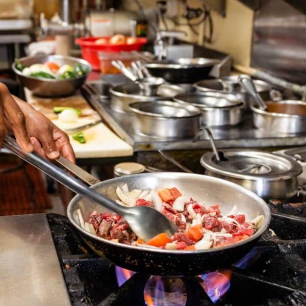 Chef's hands stirring beef, onions, and peppers in a pan over a gas flame in a commercial kitchen.