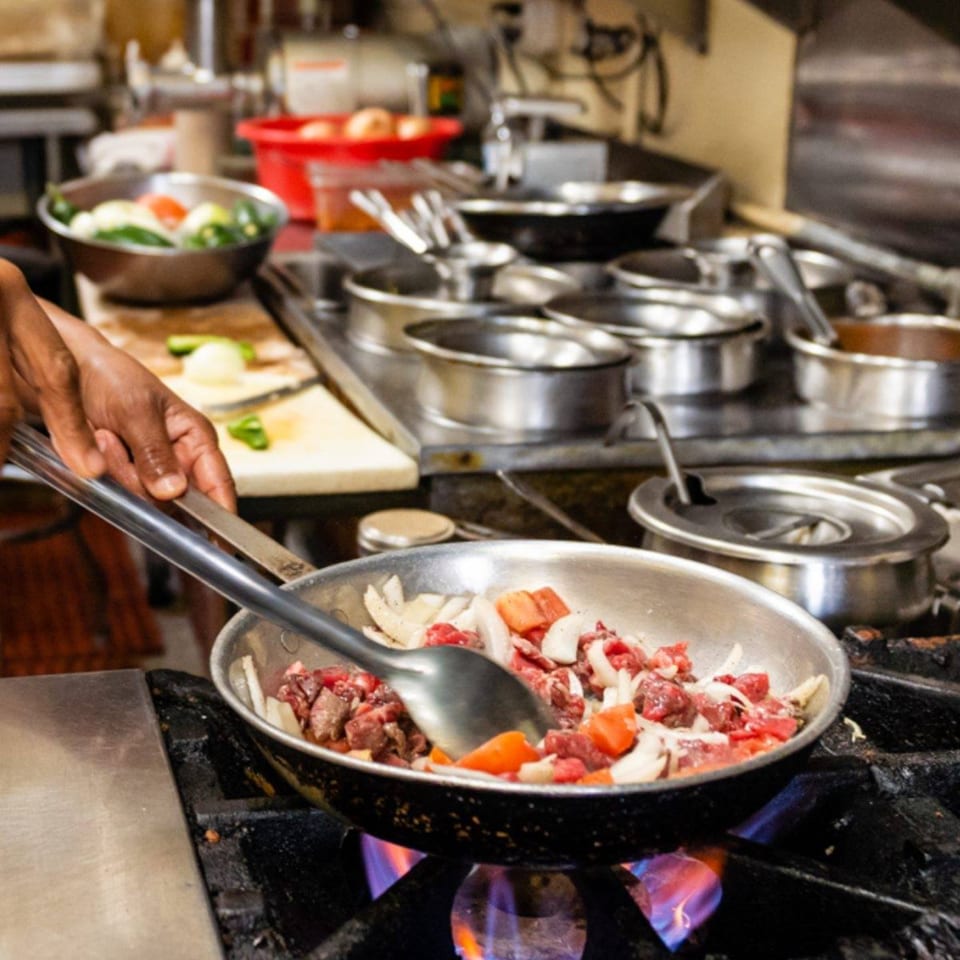 Chef's hands stirring beef, onions, and peppers in a pan over a gas flame in a commercial kitchen.